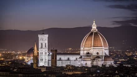 Florence skyline at night with Santa Maria del Fiore cathedral illuminated, city rooftops in the foreground, and the Apennine Mountains in the distance.