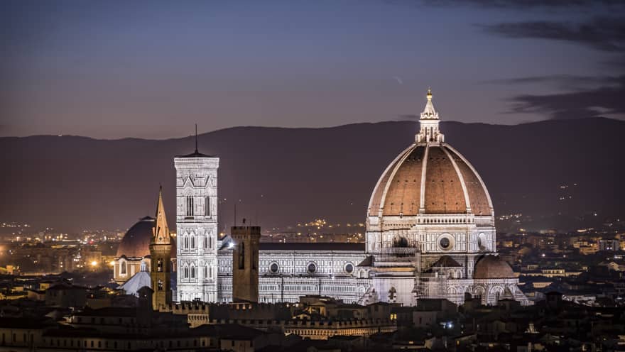 Florence skyline at night with Santa Maria del Fiore cathedral illuminated, city rooftops in the foreground, and the Apennine Mountains in the distance.